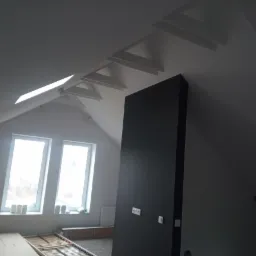 Pokój na poddaszu w trakcie wykańczania, widoczne okna dachowe, skośny sufit z elementami konstrukcyjnymi, ciemna ściana z gniazdkami i rozłożone elementy łóżka na podłodze.