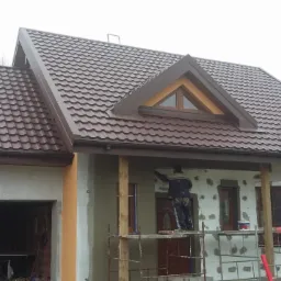 Pracownik na rusztowaniu w Łodzi tynkuje elewację domu z brązowym dachem, widoczne ocieplenie styropianem i częściowo otynkowana ściana garażu.