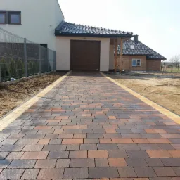 Nowa, kolorowa kostka brukowa prowadząca do garażu, obrzeża w kolorze piaskowym. W tle dom z szarą elewacją i ogrodzenie z siatki.