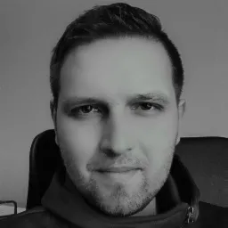 Czarno-biały portret mężczyzny z krótką brodą, skierowanego prosto w obiektyw, na neutralnym tle. Widoczne detale ubioru i fragment krzesła.