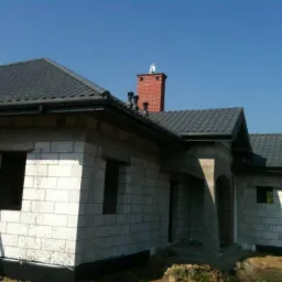 Dom w trakcie budowy z ciemnoszarym dachem z dachówki, widoczny dekarz na dachu i komin z cegły. W tle błękitne niebo.