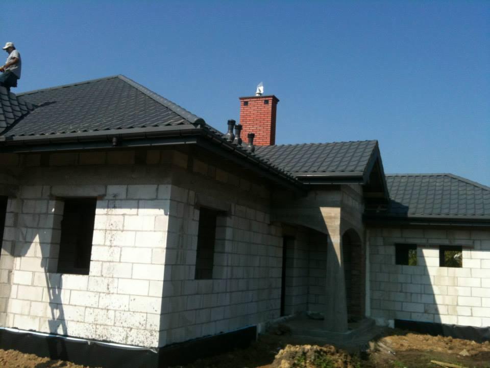 Dom w trakcie budowy z ciemnoszarym dachem z dachówki, widoczny dekarz na dachu i komin z cegły. W tle błękitne niebo.