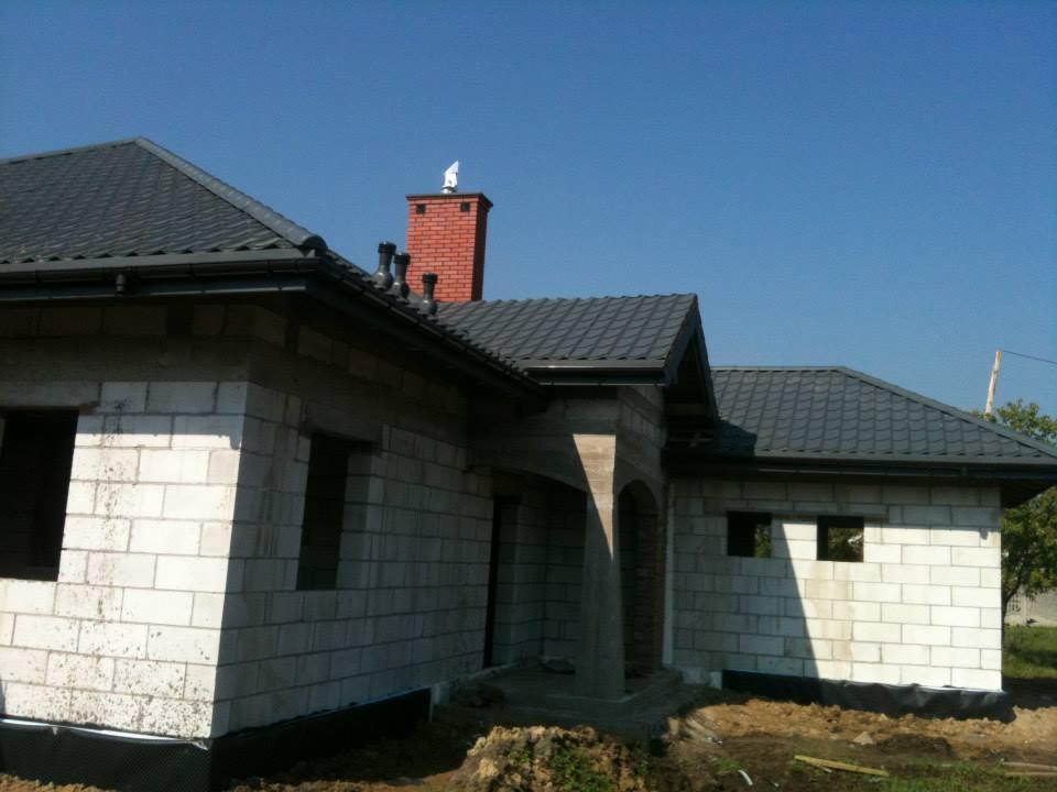 Dom w trakcie budowy z ciemnoszarą dachówką i czarnymi rynnami. Mur z białych bloczków, widoczny komin z cegły z ozdobną figurką. Błękitne niebo w tle.