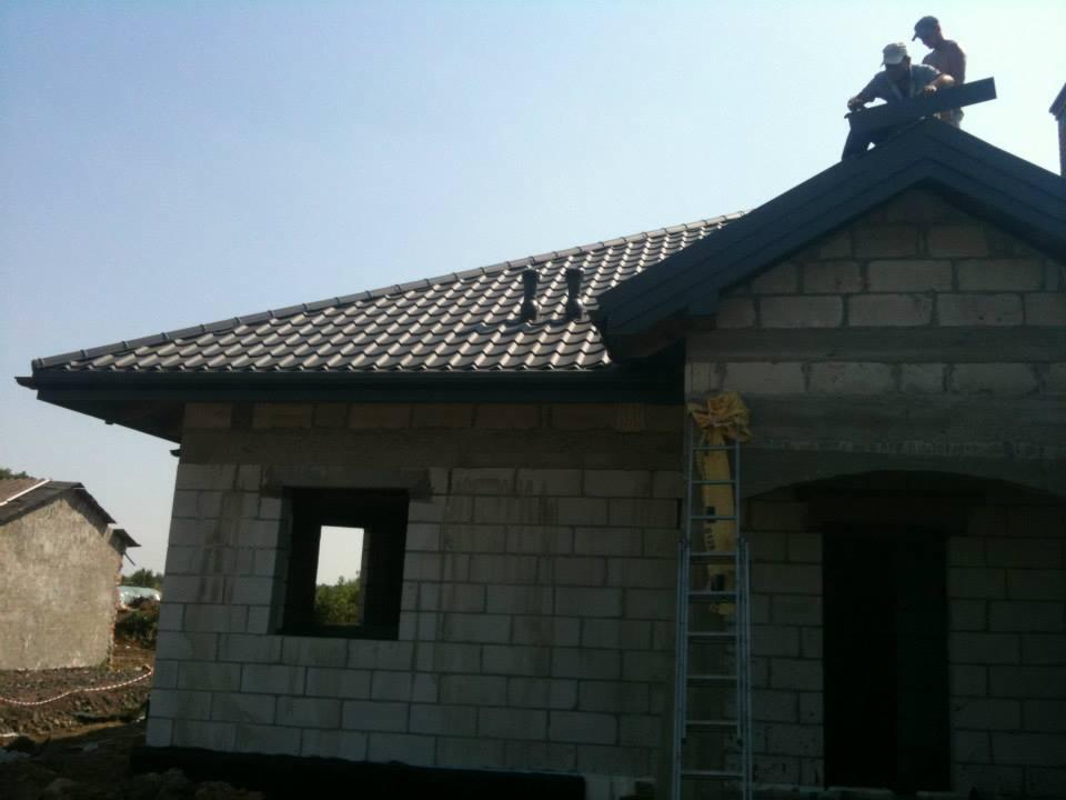 Dom w trakcie budowy z częściowo położonym dachem z dachówki ceramicznej. Na dachu dwóch dekarzy. Z boku drabina oparta o ścianę budynku.