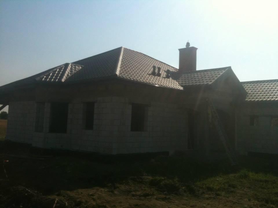 Dom w trakcie budowy z nowym, brązowym dachem z dachówki, widoczny komin i drabina oparta o ścianę. Dach pokryty w całości, elewacja surowa.