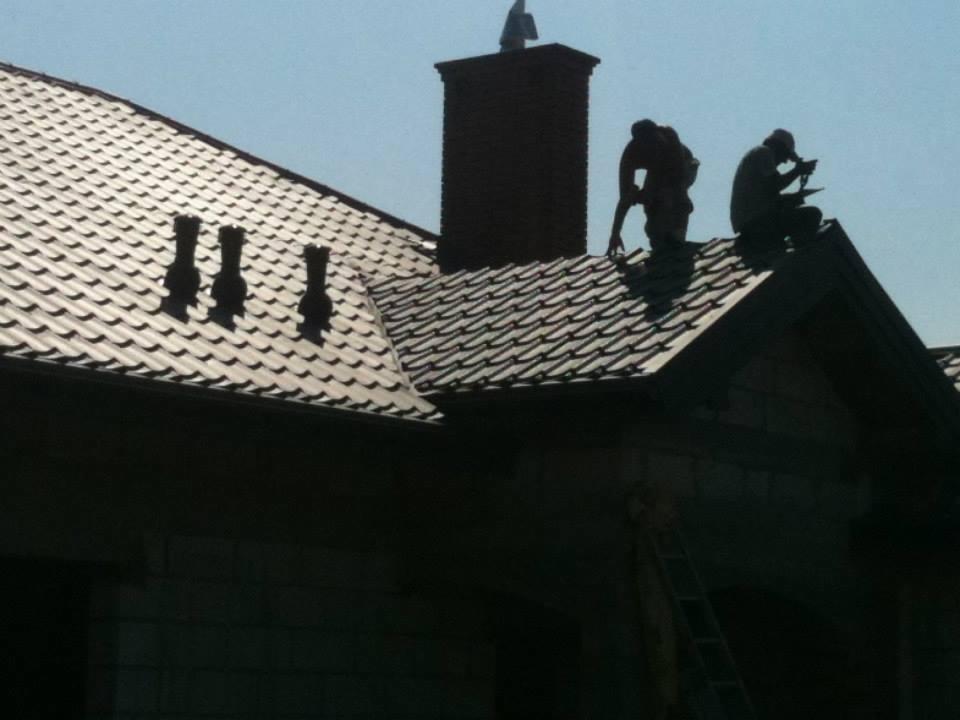 Dwóch dekarzy na dachu domu z ciemną dachówką, komin i trzy odpowietrzniki. Praca na wysokości, drabina oparta o ścianę. Silne światło słoneczne.