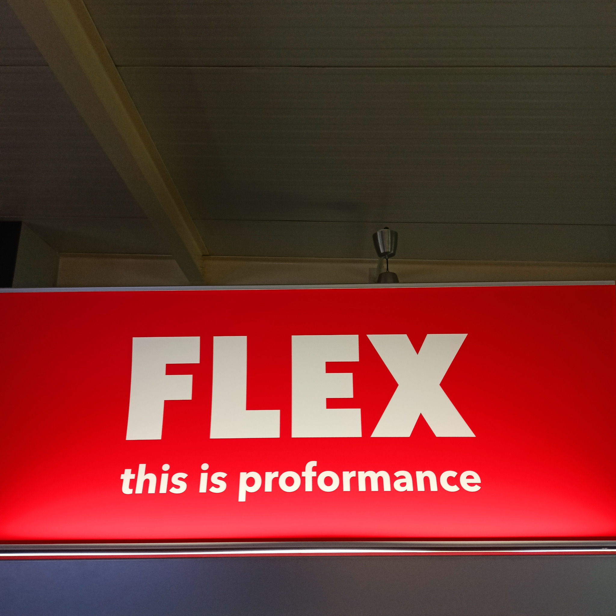 Czerwony szyld z białym napisem FLEX i hasłem 'this is proformance' pod sufitem.