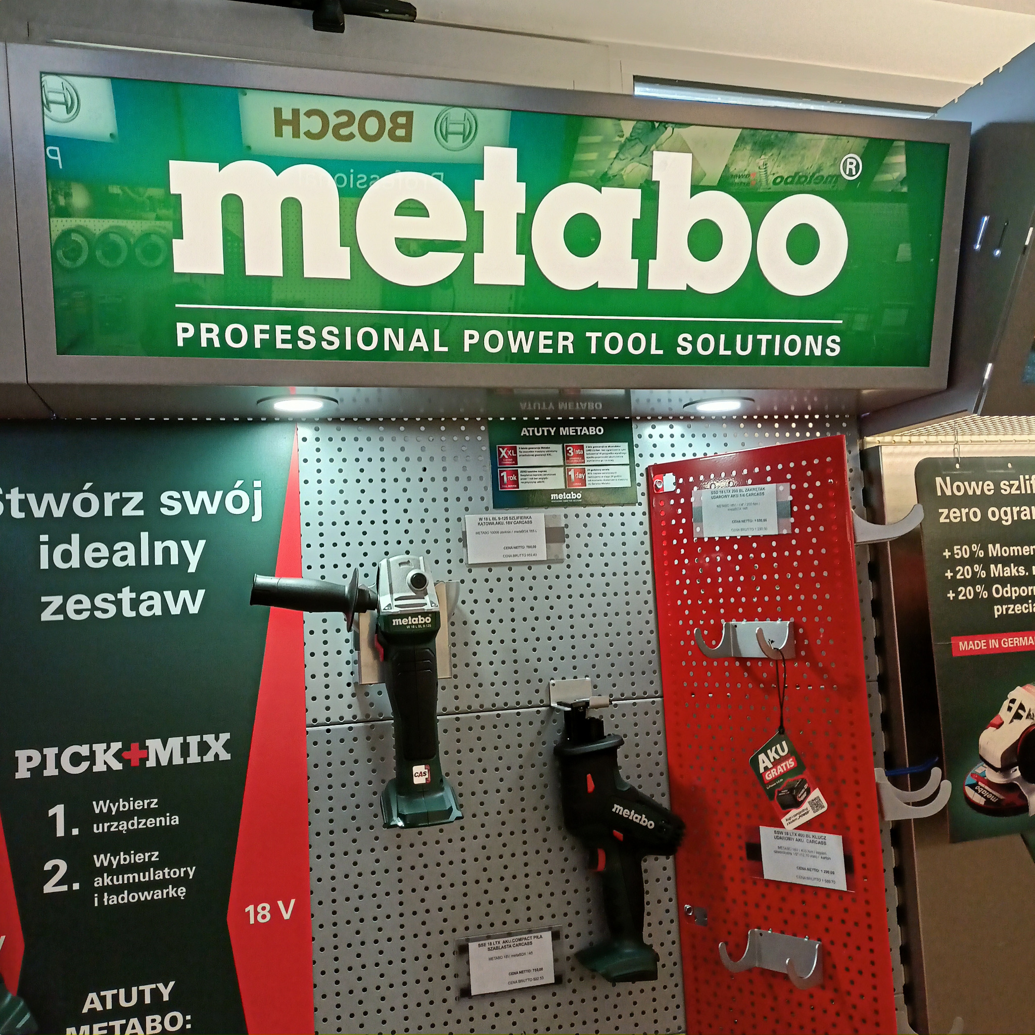 Ekspozycja elektronarzędzi Metabo w sklepie, prezentująca szlifierkę kątową, piłę szablastą i klucz udarowy na perforowanej ścianie z cenami i informacjami o promocjach.
