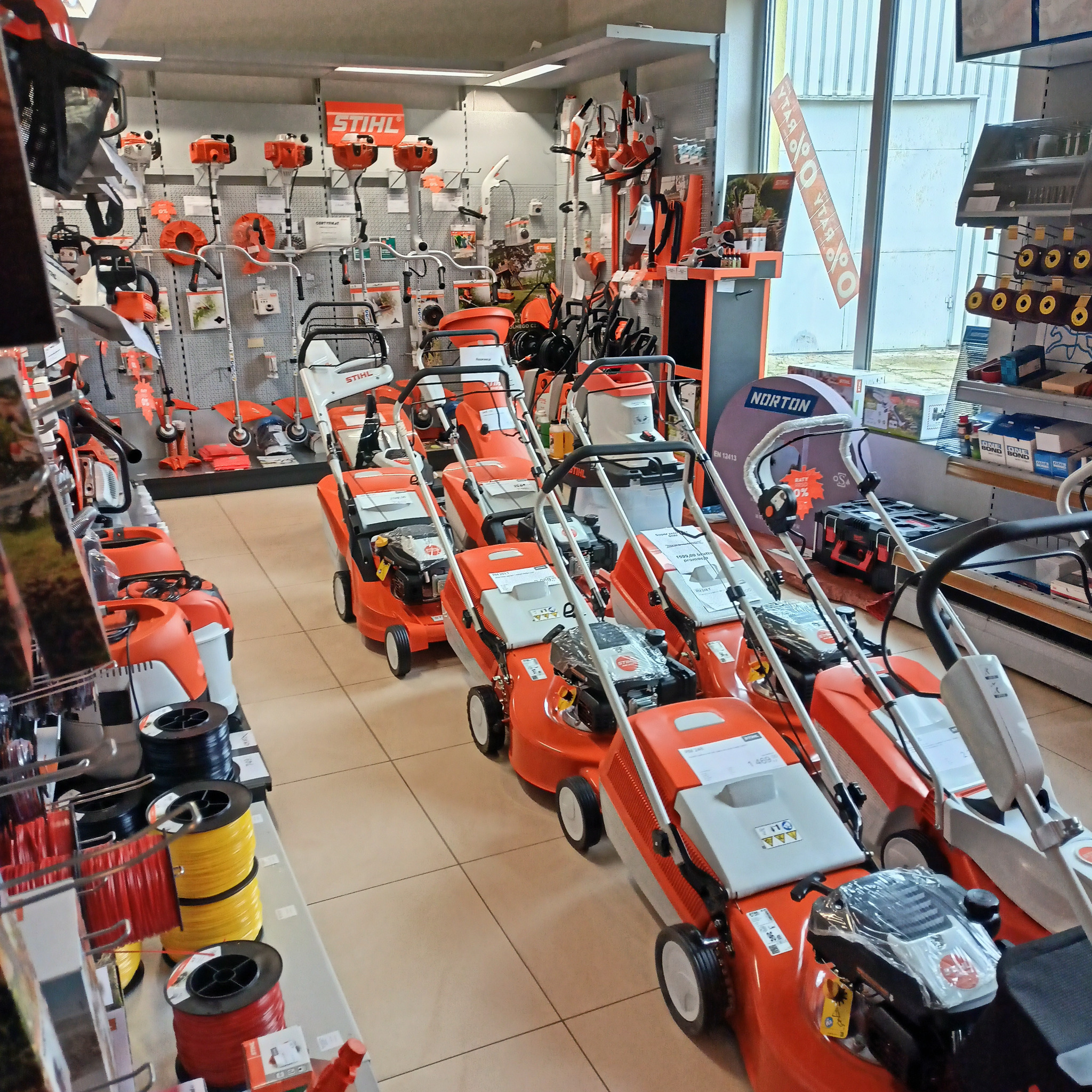 Wnętrze sklepu z maszynami ogrodniczymi, widok na rzędy nowych kosiarek Stihl w kolorze pomarańczowo-białym, na ścianach ekspozycja kos spalinowych i innych urządzeń Stihl, w tle okno z reklamą.
