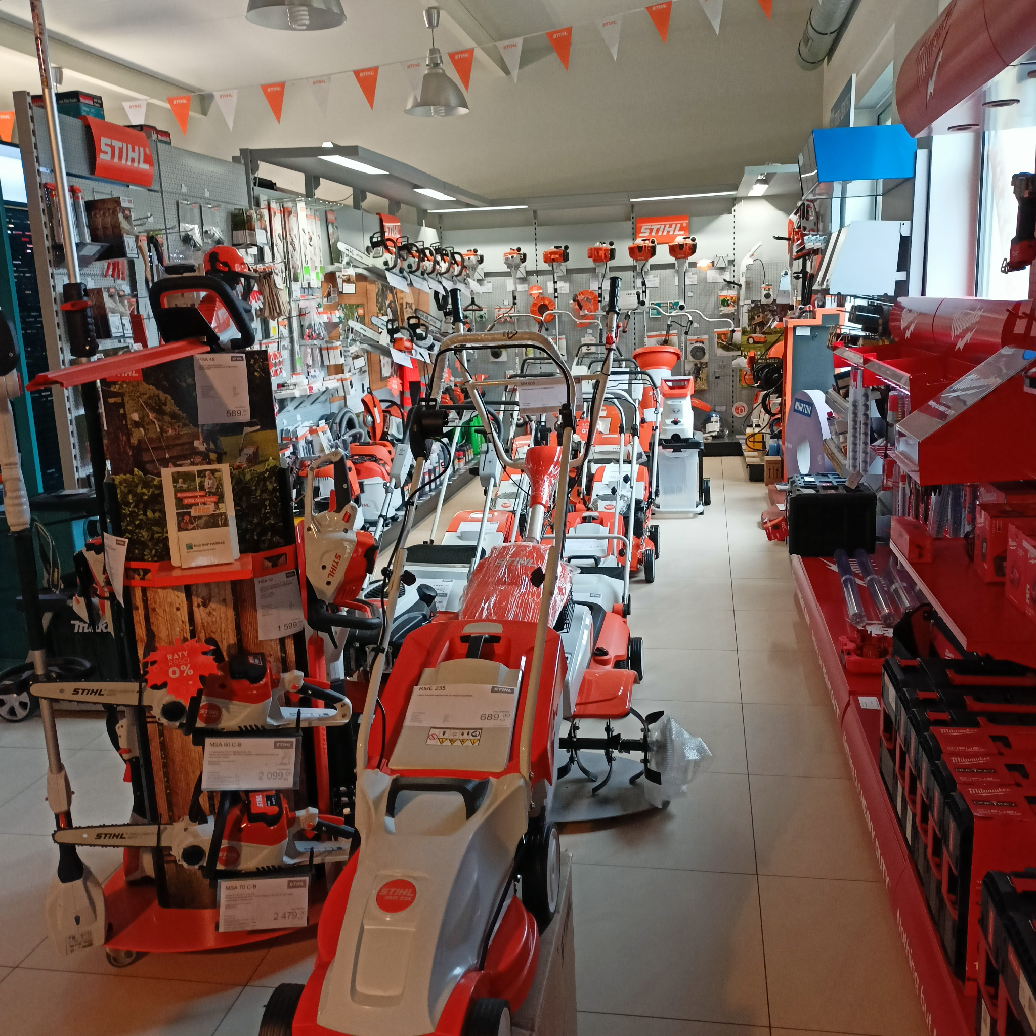 Wnętrze sklepu z narzędziami ogrodniczymi Stihl, prezentujące szeroki asortyment kosiarek, glebogryzarek i pilarek łańcuchowych na półkach i ekspozytorach, z dekoracyjnymi proporczykami marki...