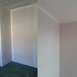 Pokój po remoncie: ściany pomalowane na jasny kolor, widoczne listwy przypodłogowe i sufitowe, na podłodze zielona powłoka. Widok na fragment pomieszczenia.