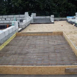 Zbrojenie fundamentu w drewnianym szalunku, widoczne bloczki betonowe i piasek. Etap budowy domu jednorodzinnego, zieleń w tle.