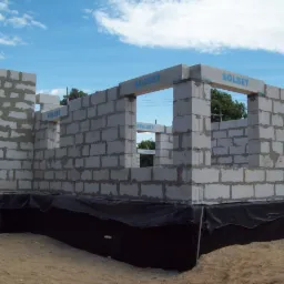 Wznoszenie ścian budynku z betonu komórkowego Solbet. Widoczne fragmenty ścian i otwory okienne. Fundament zabezpieczony folią hydroizolacyjną. Błękitne niebo w tle.