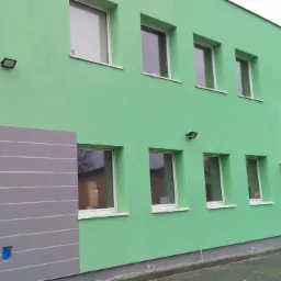 Budynek o zielonej elewacji z szarym, dekoracyjnym fragmentem narożnika. Widoczne okna z białymi ramami, oświetlenie zewnętrzne i wejście z zadaszeniem.