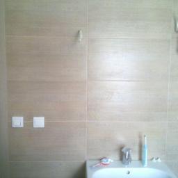 Przedsiębiorstwo Remontowo-Budowlane Robert Nowodworski - Wykończona łazienka w odcieniach beżu w Poznaniu: płytki imitujące drewno na ścianach, biała armatura, włączniki światła i wisząca żarówka. Minimalistyczny design.