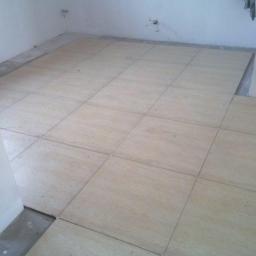 Przedsiębiorstwo Remontowo-Budowlane Robert Nowodworski - Fragment układania jasnych, kwadratowych paneli podłogowych w pomieszczeniu z poziomnicą opartą o ścianę. Widoczne niedokończone fragmenty podłoża.