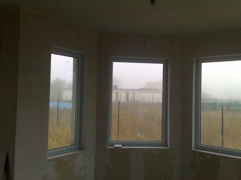 Wnętrze surowego pokoju z trzema oknami PCV, przez które widać zamglony krajobraz z budynkiem w tle. Ściany wymagają wykończenia, widoczna żarówka na suficie.