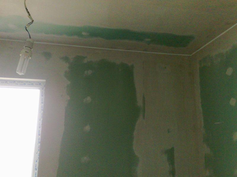 Ściana po szpachlowaniu, widoczne nierówności i zielone plamy. W rogu sufitu listwa, a z góry zwisa żarówka energooszczędna. Okno po lewej stronie.