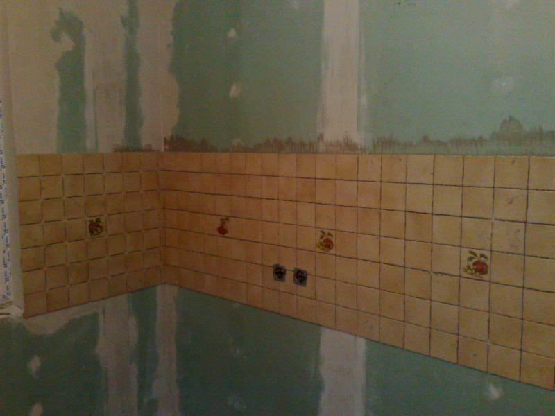 Ściana w kuchni z beżowymi płytkami z motywem kwiatowym, częściowo ułożona. Widoczne gniazdka elektryczne i surowe ściany w tle. Podłoga zabezpieczona folią.