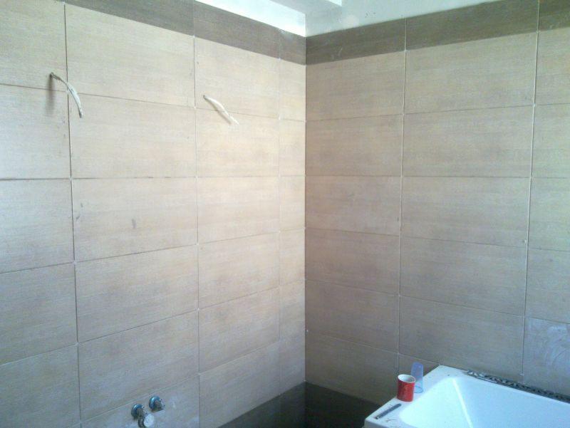 Wyłożona beżowymi płytkami łazienka z wystającymi przewodami i fragmentem wanny. Widoczna szara listwa dekoracyjna pod sufitem.