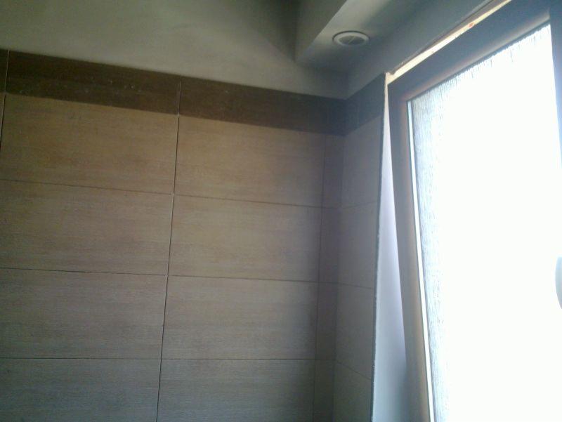 Wykończona ściana w łazience z beżowymi płytkami i ciemniejszym pasem na górze, widoczne okno z jasnym światłem dziennym i wbudowane oświetlenie sufitowe.