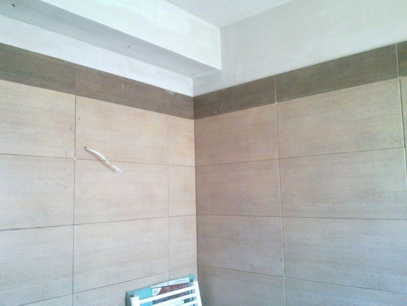 Wykończona ściana łazienki z beżowymi płytkami i brązowym pasem dekoracyjnym u góry. Widoczny wystający kabel i fragment drabinki łazienkowej.