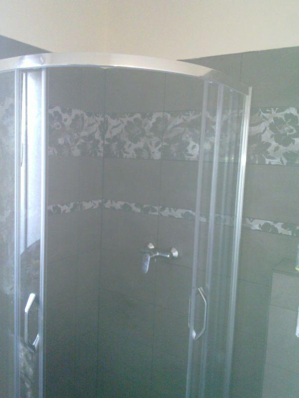 Szklana kabina prysznicowa z chromowanymi detalami na tle ciemnych, wzorzystych płytek w łazience. Widoczna bateria i uchwyty. Wykończona łazienka po remoncie.