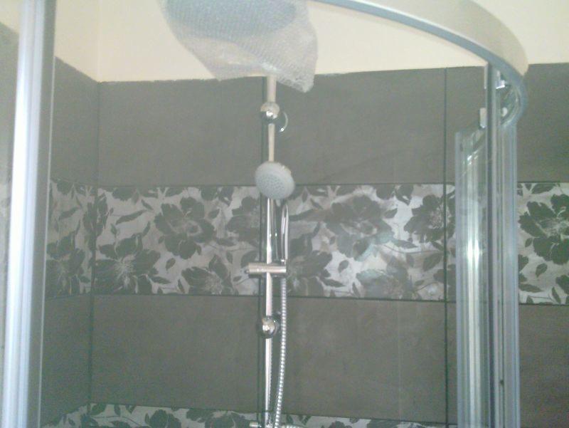 Szara kabina prysznicowa z kwiatowym motywem na płytkach. Nowa słuchawka prysznicowa z folią ochronną. Widok na fragment łazienki po remoncie.
