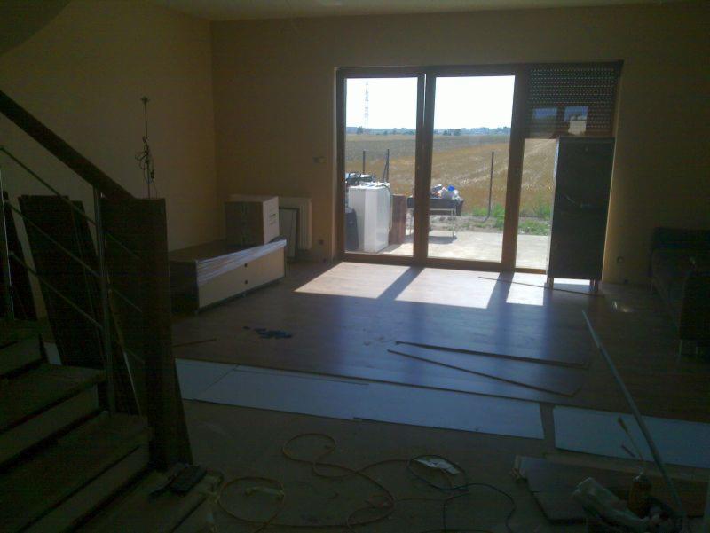 Salon w trakcie remontu: częściowo ułożona drewniana podłoga, widoczne panele i kable, duże okno z widokiem na pole. Meble zabezpieczone folią.