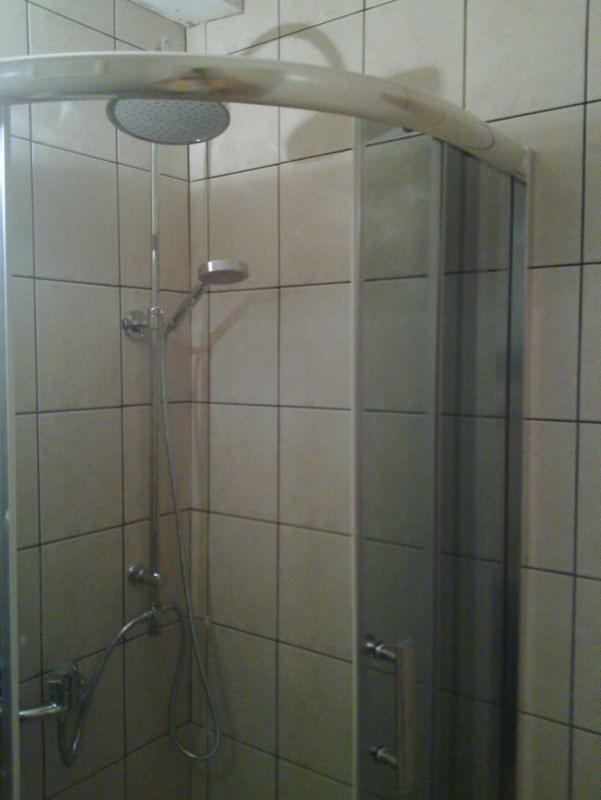 Kabina prysznicowa z deszczownicą i szklanymi drzwiami w łazience wyłożonej beżowymi, kwadratowymi płytkami ceramicznymi. Widoczne elementy armatury.