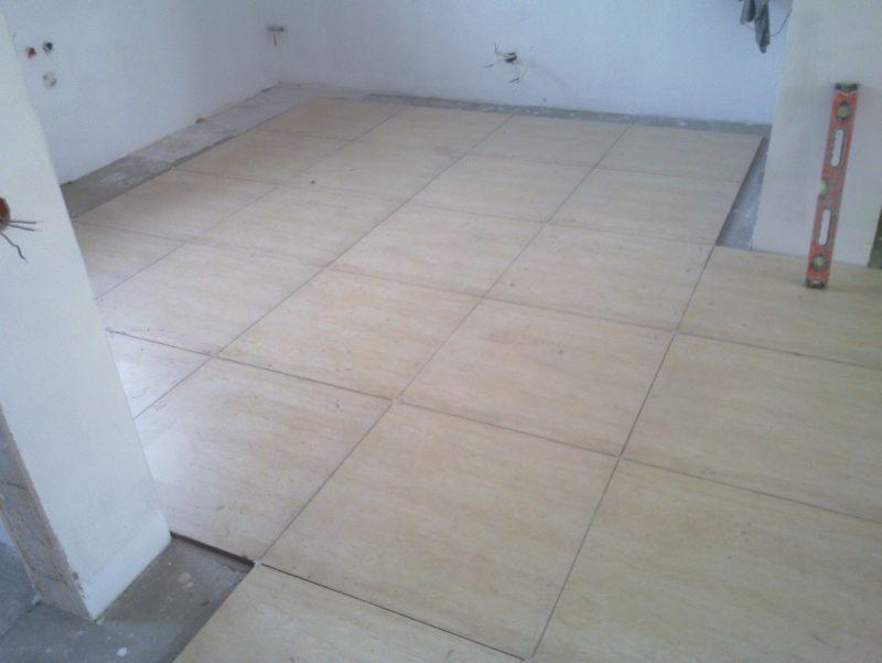 Fragment układania jasnych, kwadratowych paneli podłogowych w pomieszczeniu z poziomnicą opartą o ścianę. Widoczne niedokończone fragmenty podłoża.