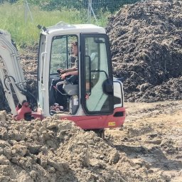 Usługi sprzętem ogrodniczym Daniel Juszczak - Operator w miniładowarce Takeuchi, praca w błotnistym terenie. Widoczna sterta ziemi i częściowo wykop. Praca w trakcie niwelacji terenu.