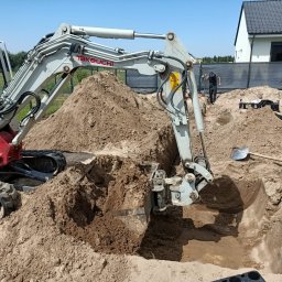 Usługi sprzętem ogrodniczym Daniel Juszczak - Mini koparka Takeuchi w trakcie wykopu pod fundamenty domu jednorodzinnego. W tle widoczny pracownik i fragment budynku.