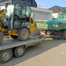 Usługi sprzętem ogrodniczym Daniel Juszczak - Żółta miniładowarka Wacker Neuson WL25 na niskopodwoziowej przyczepie za zieloną ciężarówką MAN, w tle nowoczesny dom z czarnym dachem i białą elewacją.