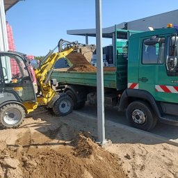 Usługi sprzętem ogrodniczym Daniel Juszczak - Żółta miniładowarka Wacker Neuson nabiera piasek i wysypuje go na pakę zielonej ciężarówki MAN na tle budynku o jasnych ścianach i niebieskiego nieba.