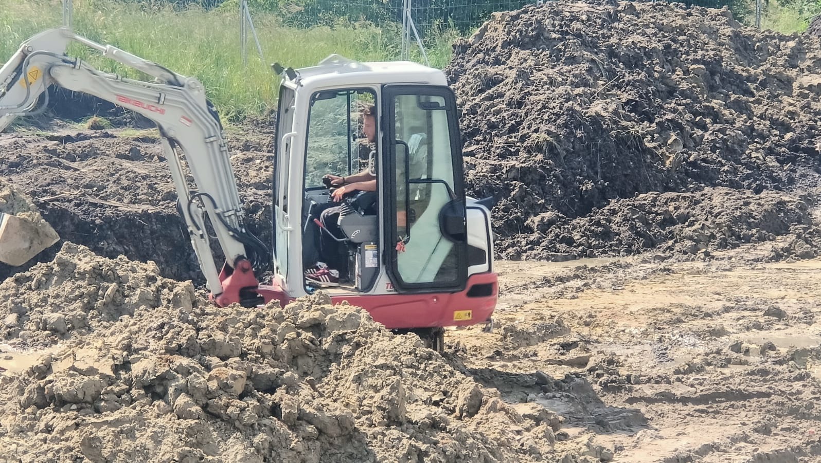 Operator w miniładowarce Takeuchi, praca w błotnistym terenie. Widoczna sterta ziemi i częściowo wykop. Praca w trakcie niwelacji terenu.