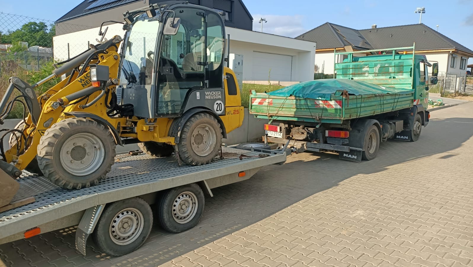 Żółta miniładowarka Wacker Neuson WL25 na niskopodwoziowej przyczepie za zieloną ciężarówką MAN, w tle nowoczesny dom z czarnym dachem i białą elewacją.