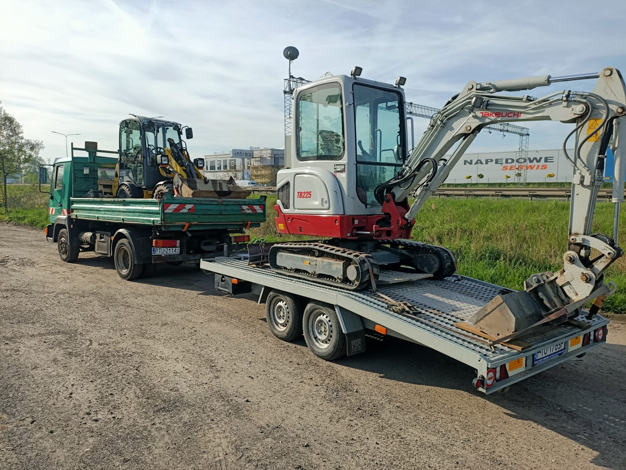 Transport dwóch mini koparek: biało-czerwonej Takeuchi TB225 na niskopodwoziowej przyczepie i żółtej Wacker Neuson na zielonej ciężarówce MAN, na żwirowej drodze.