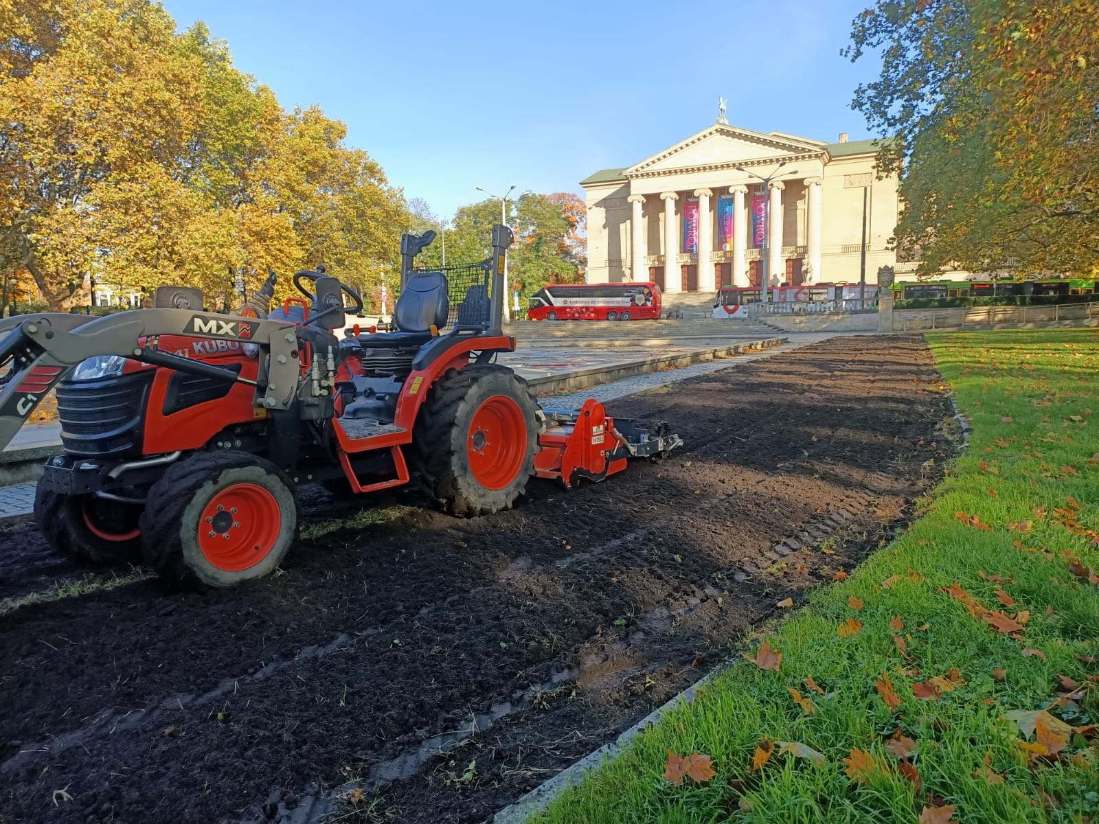 Czerwony traktor Kubota MX41 z glebogryzarką przygotowuje teren pod trawnik na tle klasycystycznego budynku i jesiennych drzew w słoneczny dzień.
