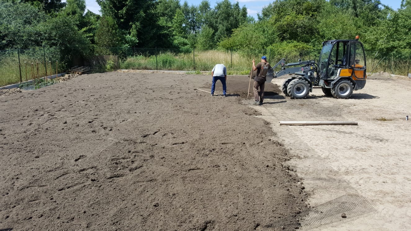 Przygotowanie terenu pod trawnik: dwaj pracownicy wyrównują ziemię grabiami, w tle miniładowarka Giant i zieleń. Część terenu wyłożona siatką wzmacniającą.
