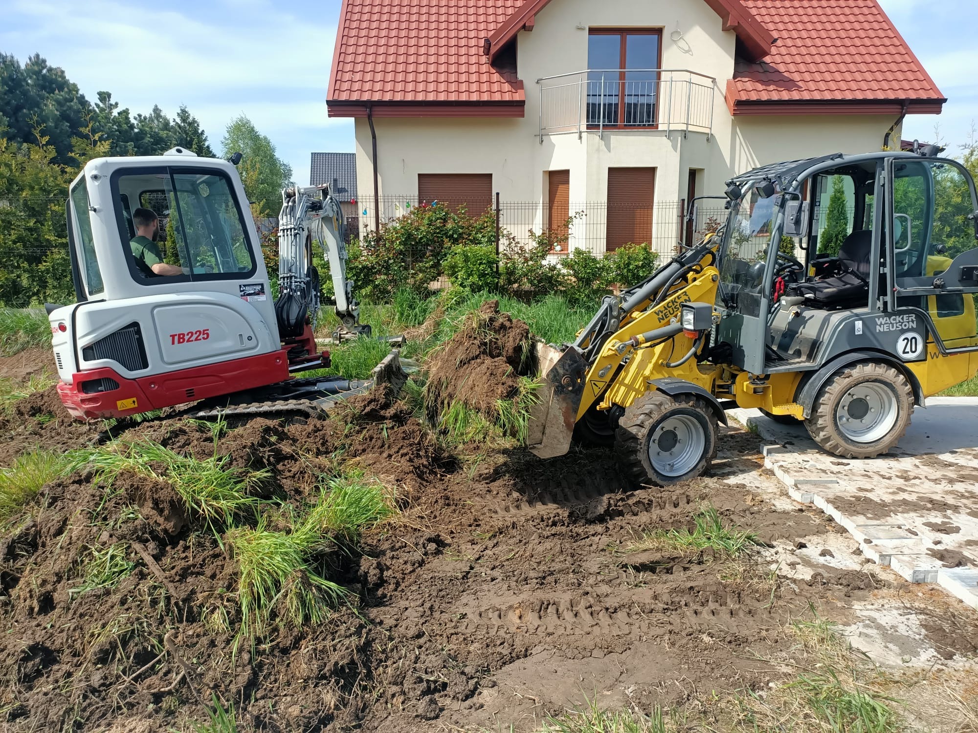 Żółta miniładowarka i biało-czerwona koparka na placu budowy przed domem. Wykopane ziemia i ślady opon. Prace ziemne w toku, maszyny Wacker Neuson.