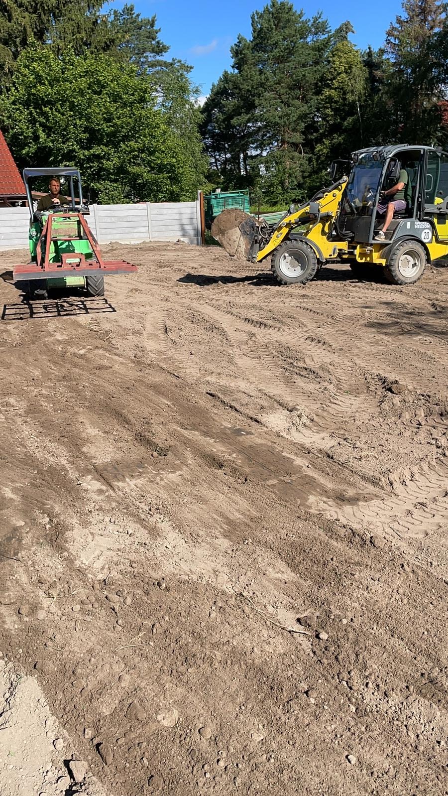 Żółta miniładowarka i zielony traktor z broną wyrównują teren. W tle drzewa i szary płot. Prace ziemne w toku, przygotowanie podłoża pod trawnik.