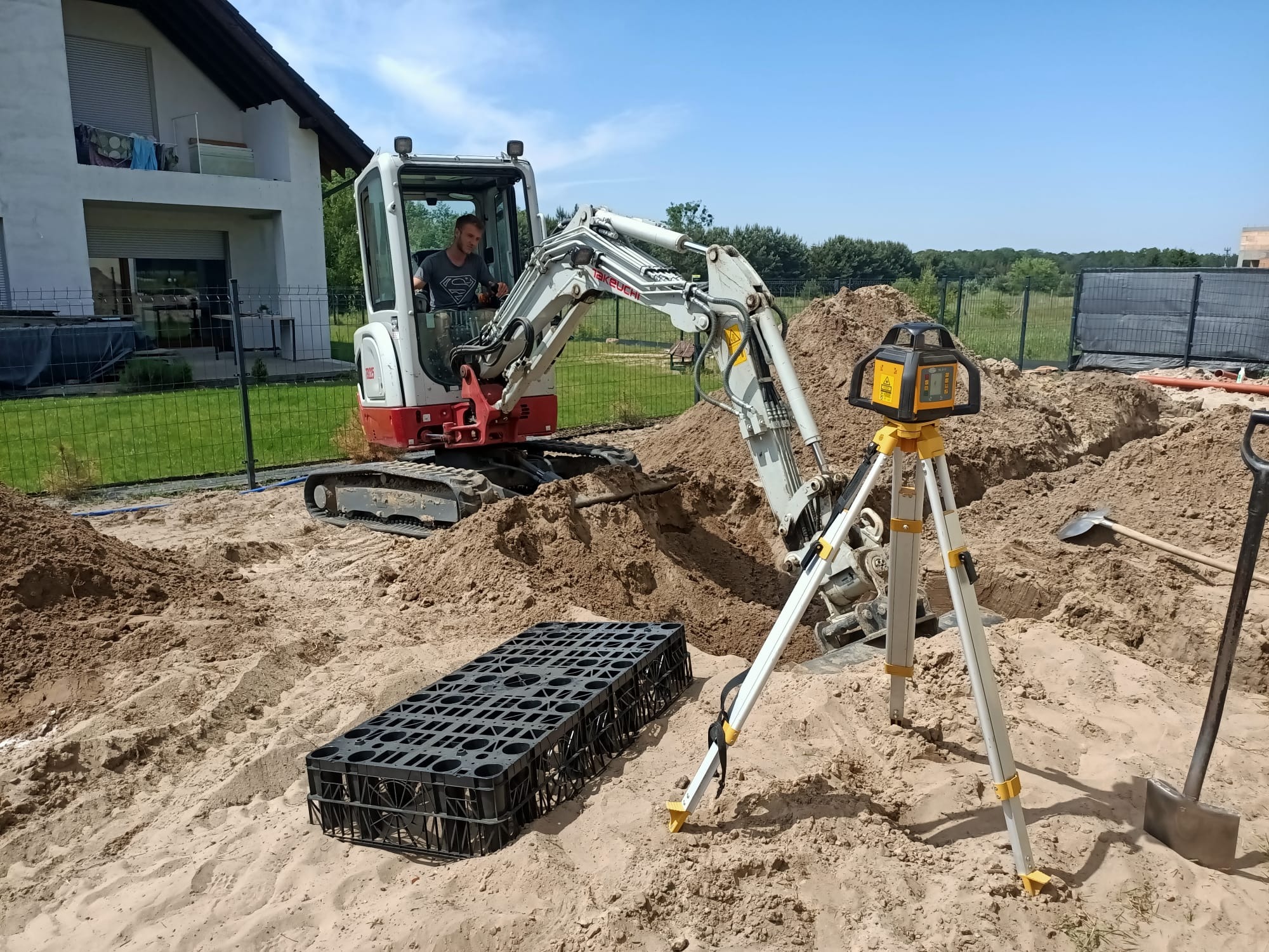 Mini koparka Takeuchi w wykopie, operator w kabinie, obok czarna skrzynka rozsączająca i niwelator laserowy na statywie. W tle dom i zieleń.