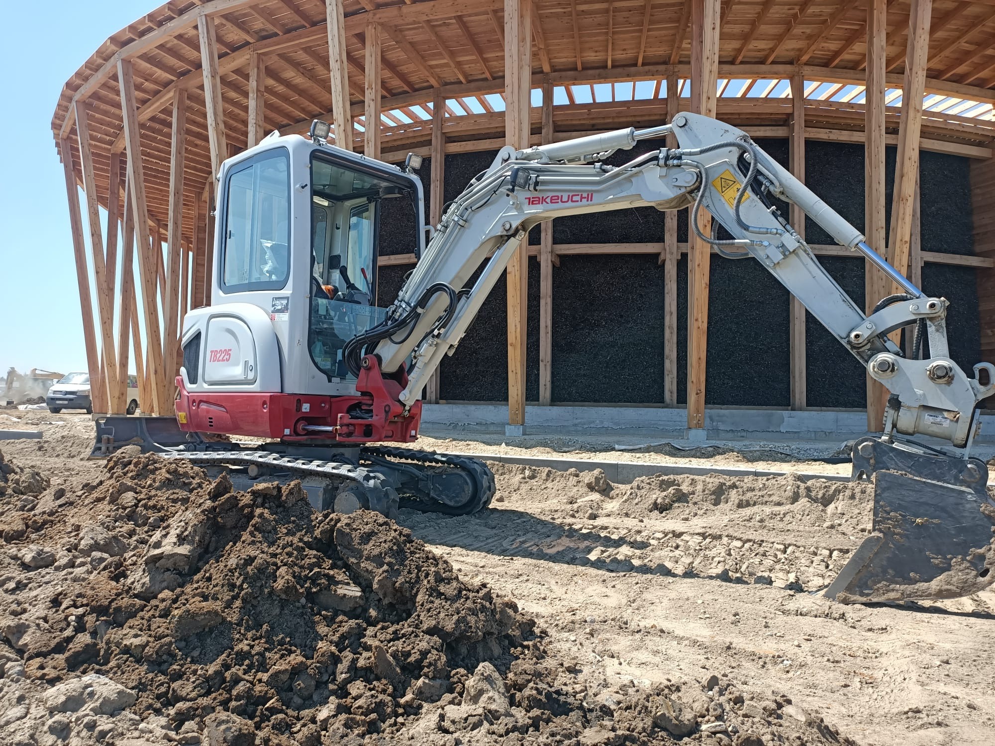 Mini koparka Takeuchi TB225 na placu budowy, z drewnianą konstrukcją w tle. Wykopane ziemia i piasek, przygotowanie terenu pod budowę.