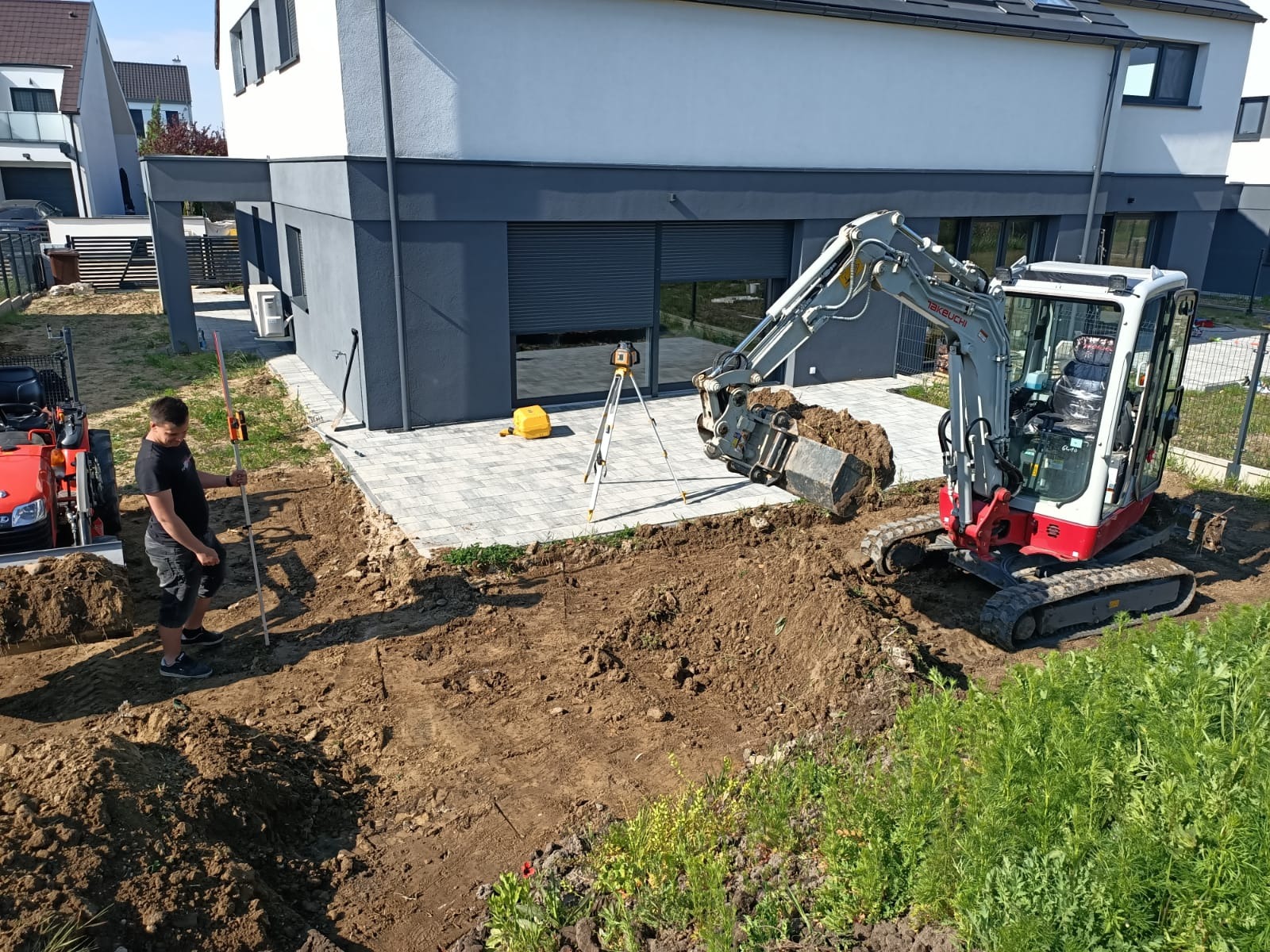 Mini koparka Takeuchi wykonuje wykop pod instalacje obok nowoczesnego domu. Pracownik z łatą niwelacyjną kontroluje głębokość. W tle poziomica laserowa.
