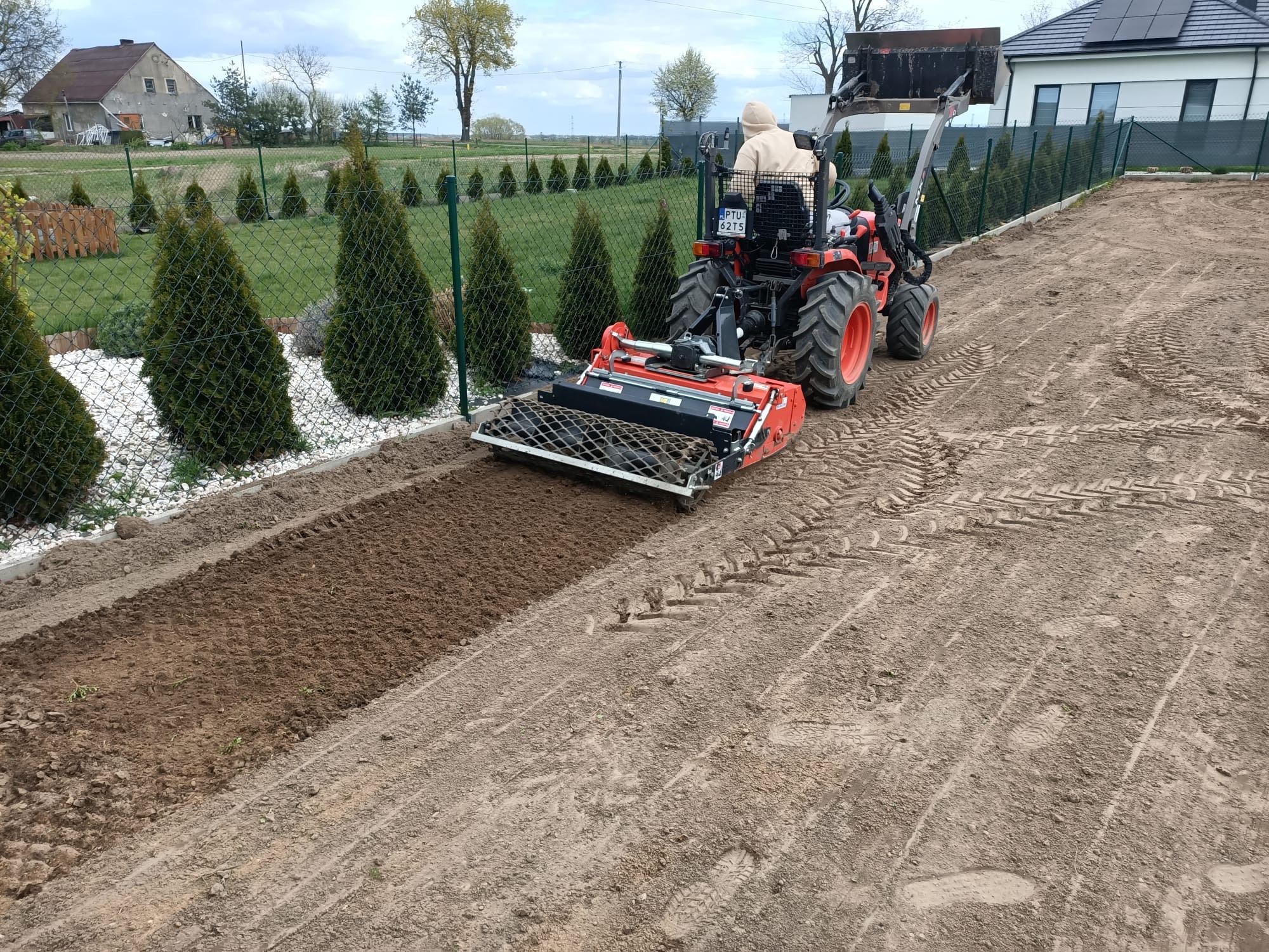 Czerwony traktor z glebogryzarką przygotowuje ziemię pod trawnik w Poznaniu. Widoczne świeżo spulchnione pasy ziemi i zielone tuje wzdłuż ogrodzenia.