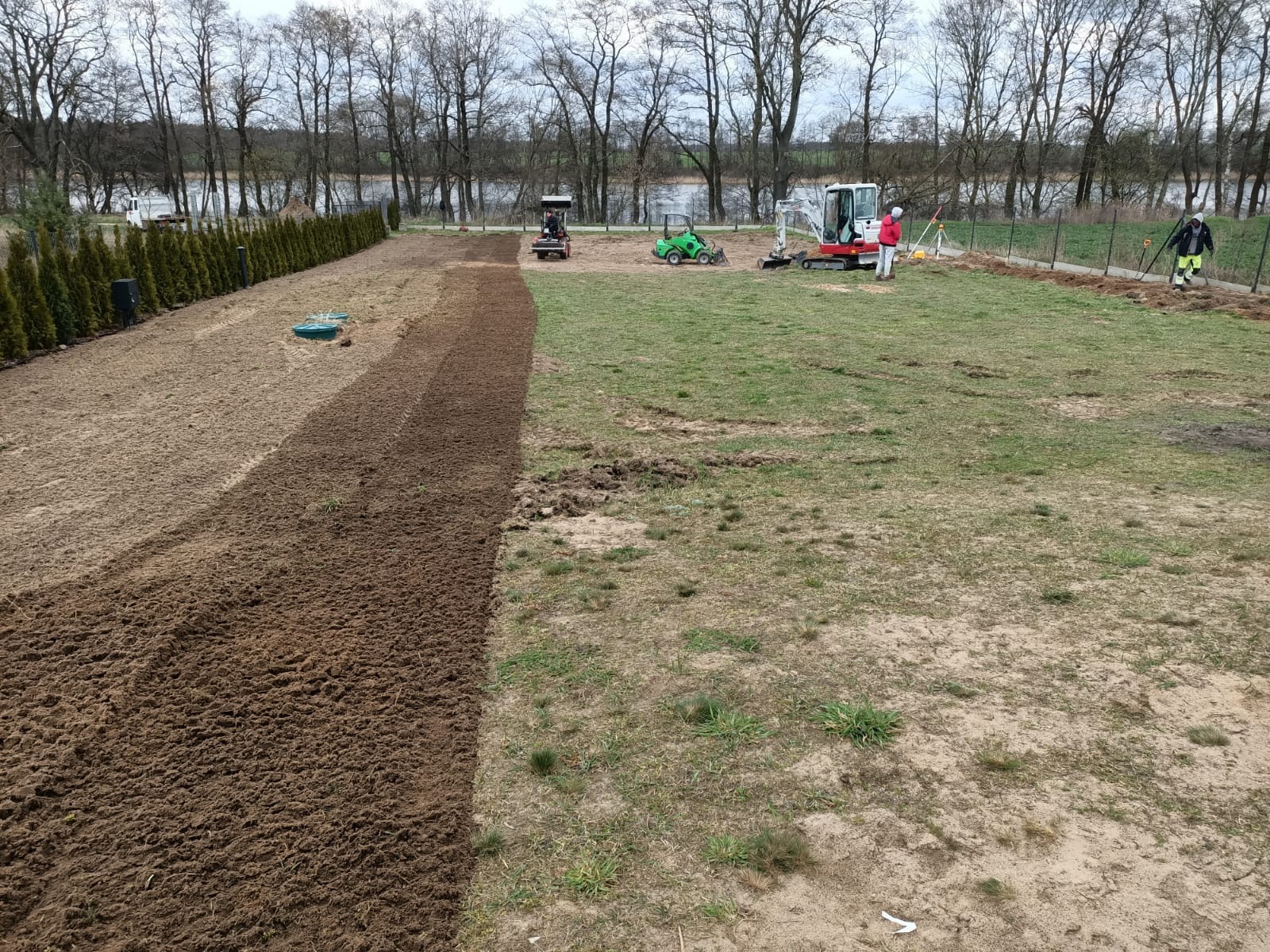 Przygotowanie terenu pod trawnik: wyrównana gleba, miniładowarki, ekipa geodetów. Częściowo przygotowany teren pod trawnik, widok na jezioro w tle.