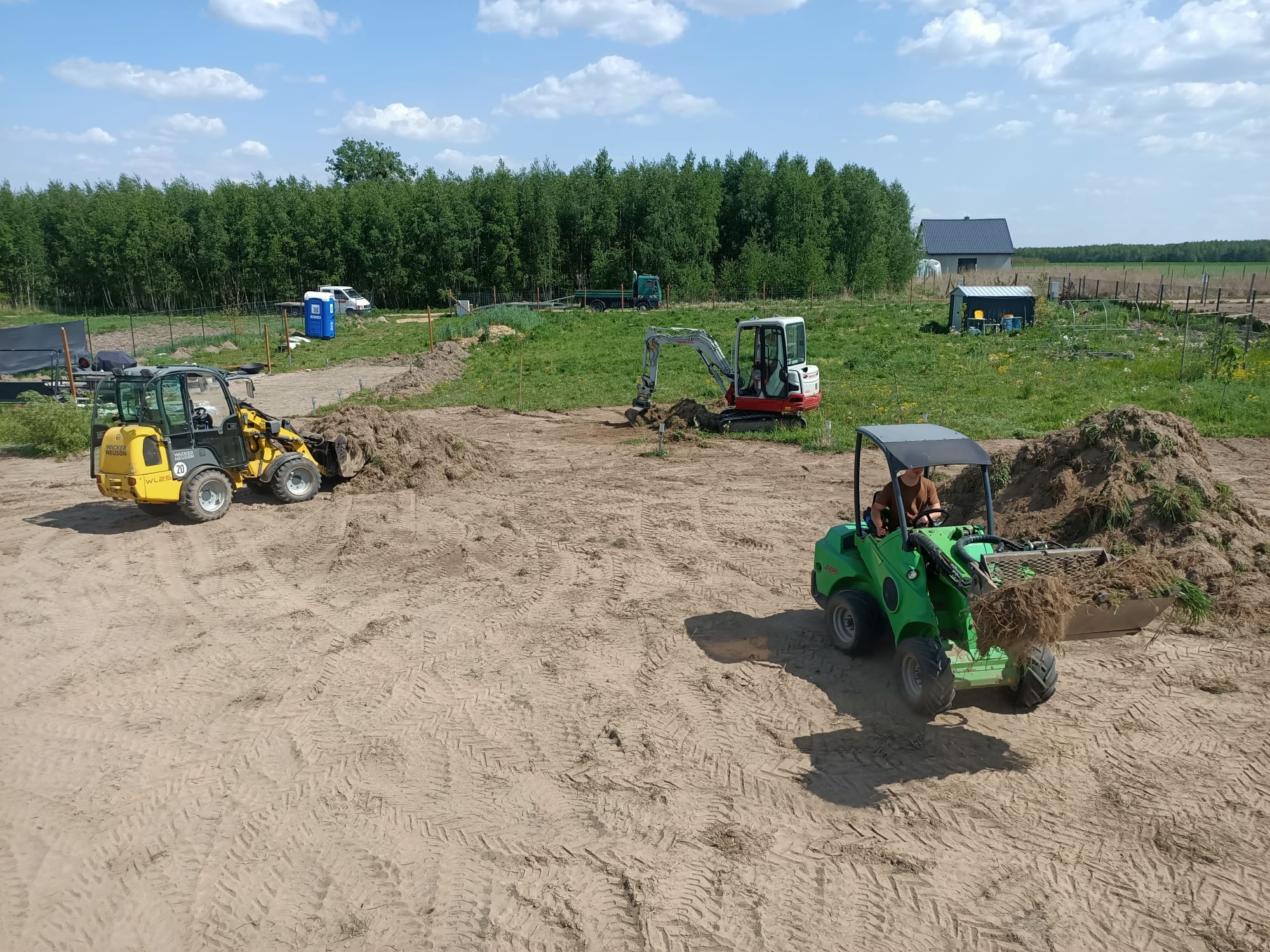 Niwelacja terenu za pomocą miniładowarek: żółtej i zielonej, oraz koparki. W tle widoczny las i budynek. Prace ziemne w toku, teren przygotowywany pod inwestycję.