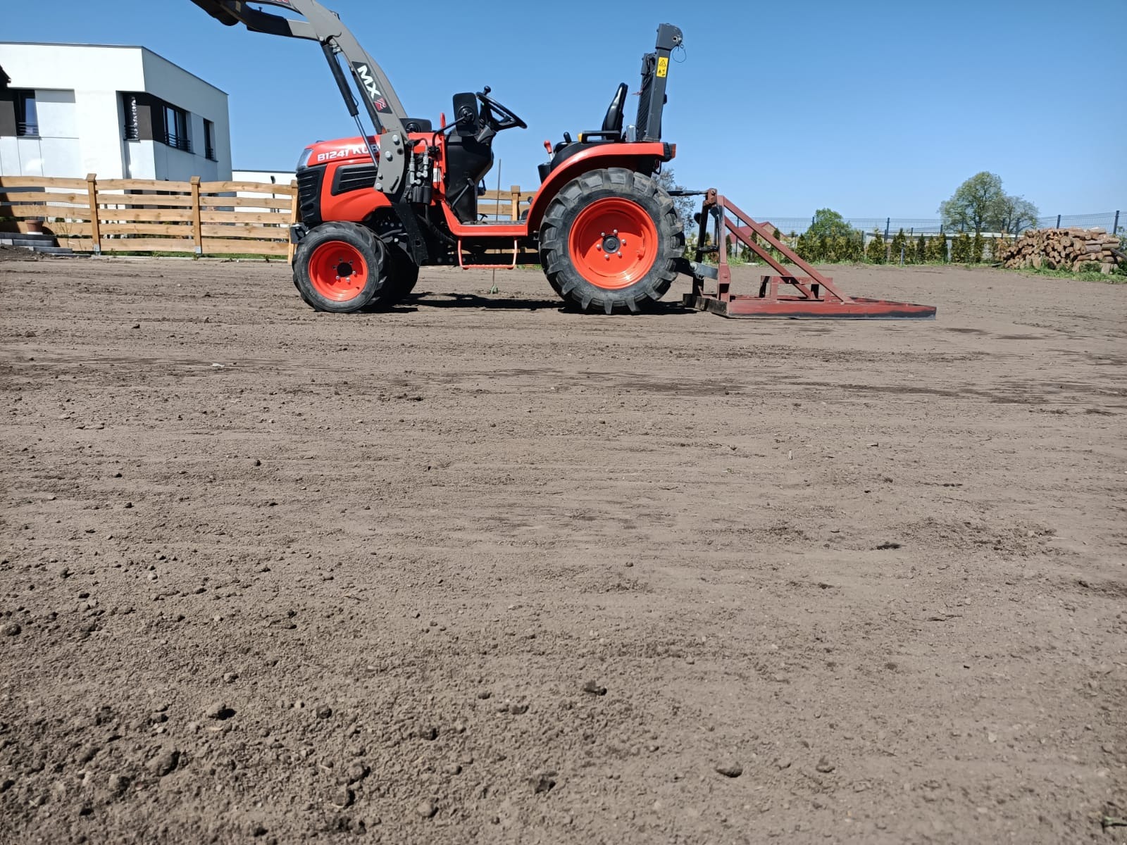 Czerwony traktor z równiarką na polu, przygotowany teren pod budowę lub ogród. W tle budynek i ogrodzenie. Błękitne niebo.