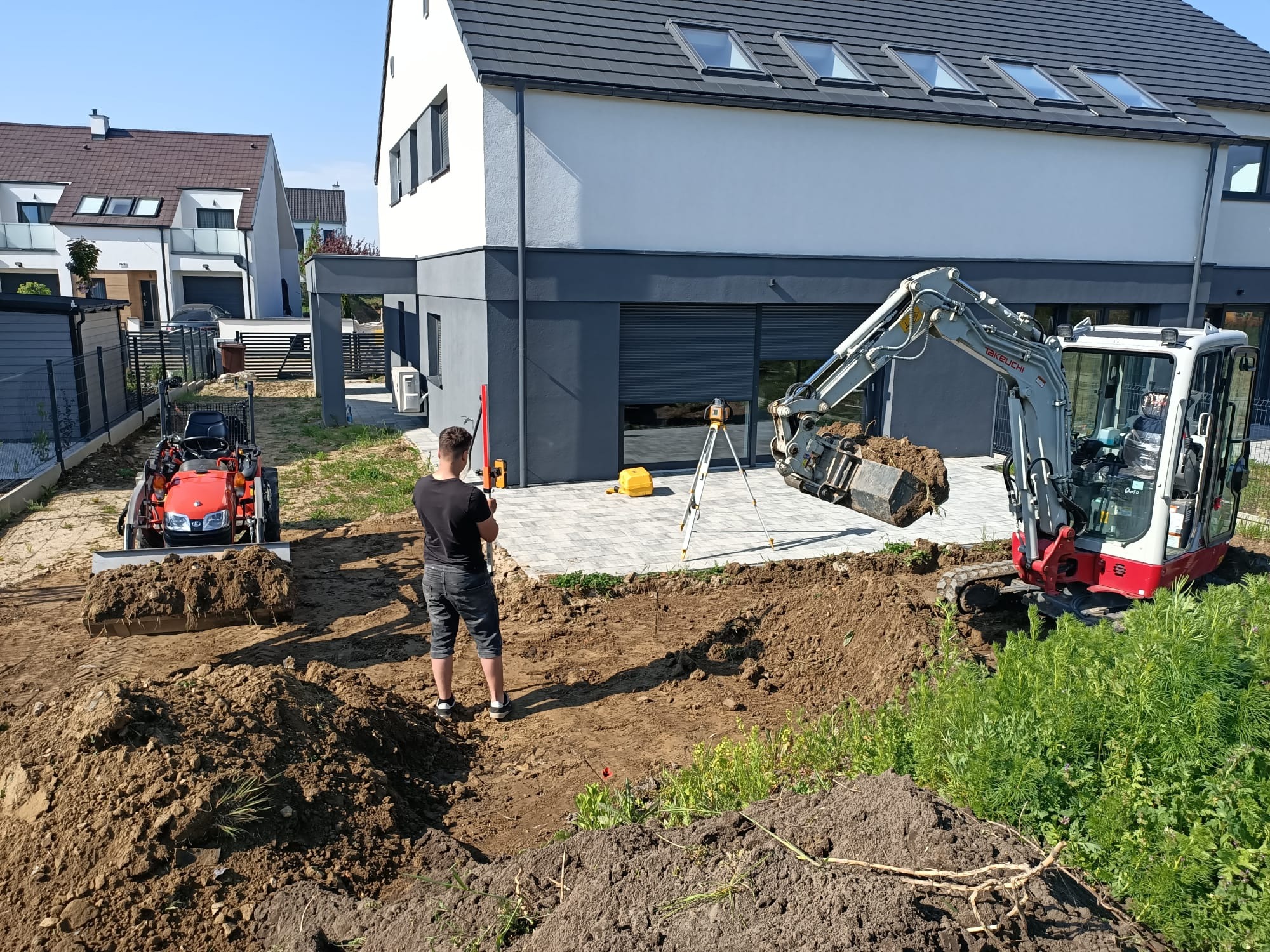 Niwelacja terenu pod budowę domu: miniładowarka, operator z niwelatorem laserowym, wykopana ziemia i dom w tle. Prace ziemne w toku, widok z perspektywy.